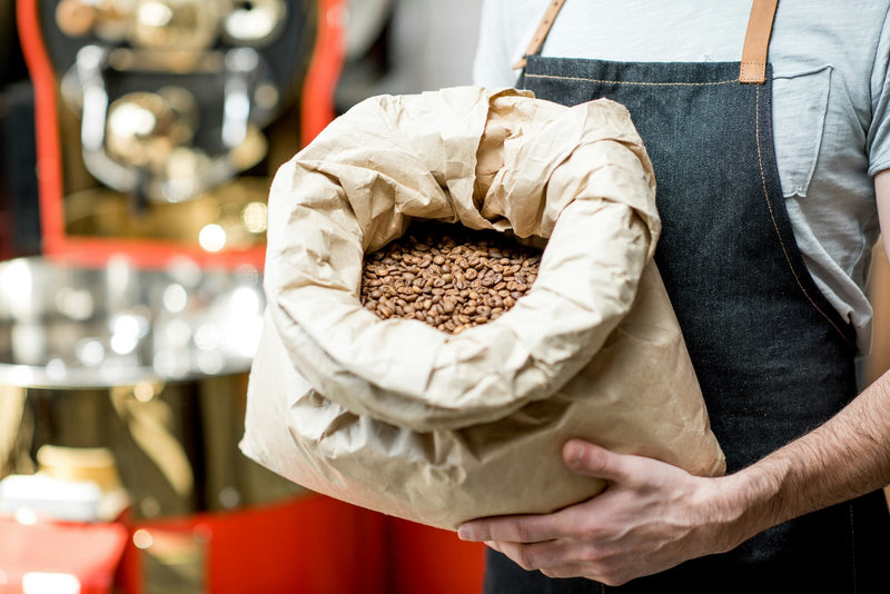 Coffee roaster holding a large paper bag of freshly roasted beans in front of a roasting machine