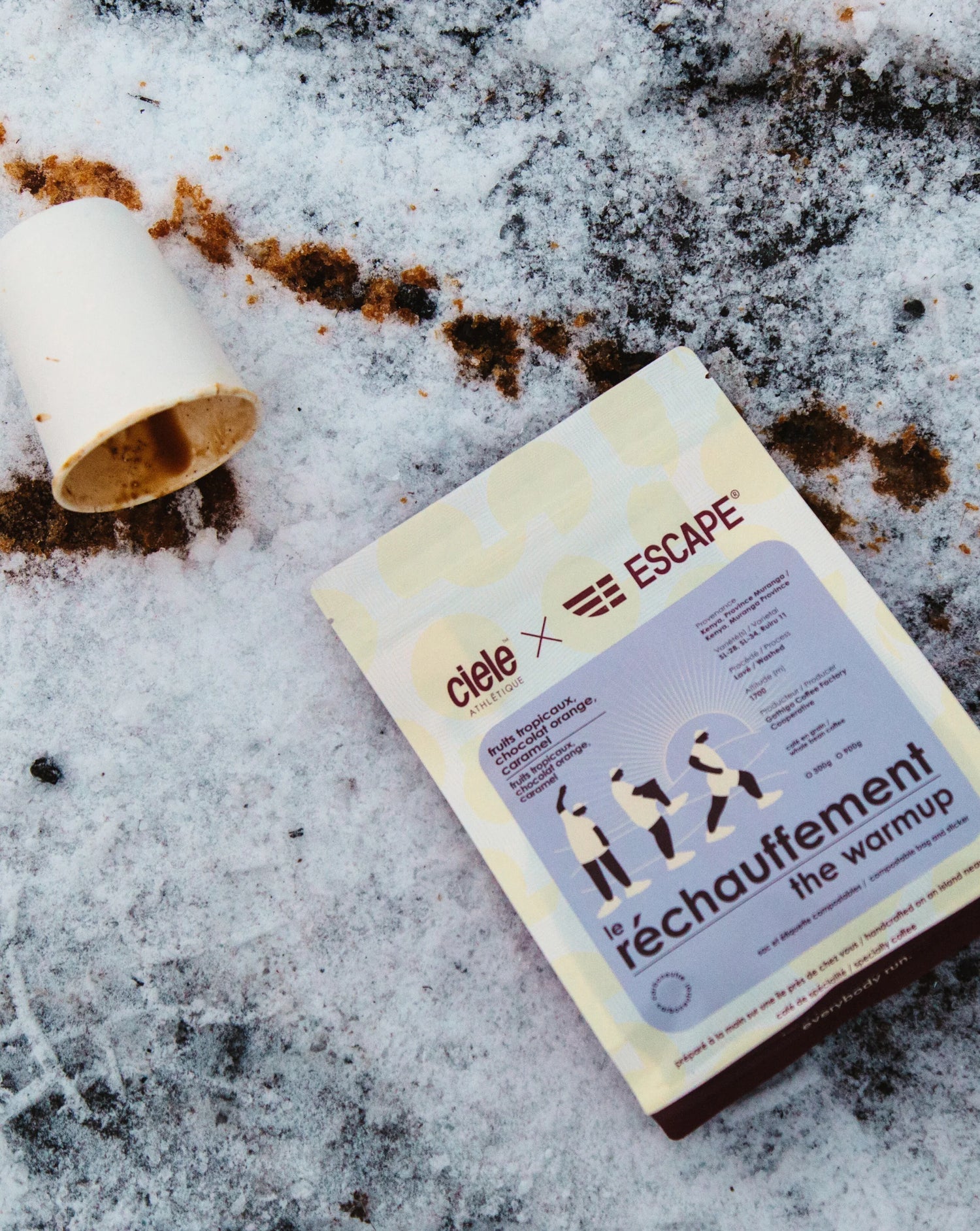 Package of Ciele x ESCAPE “Le réchauffement / The Warmup” hot chocolate mix on snow beside a tipped paper cup.