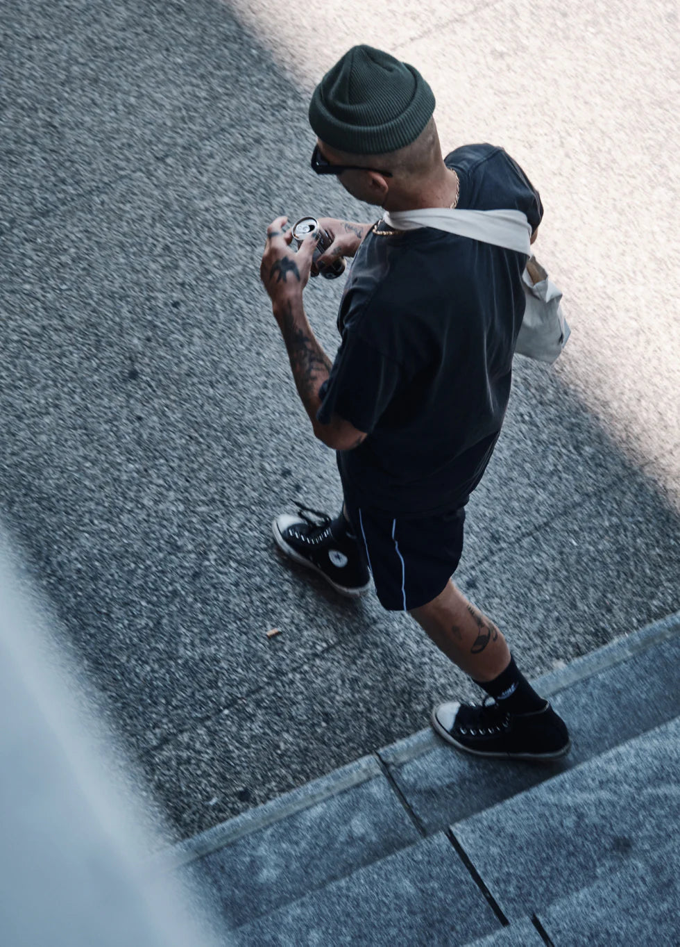 Person wearing a beanie, sunglasses, and a crossbody bag walking on a city sidewalk while holding a drink can.